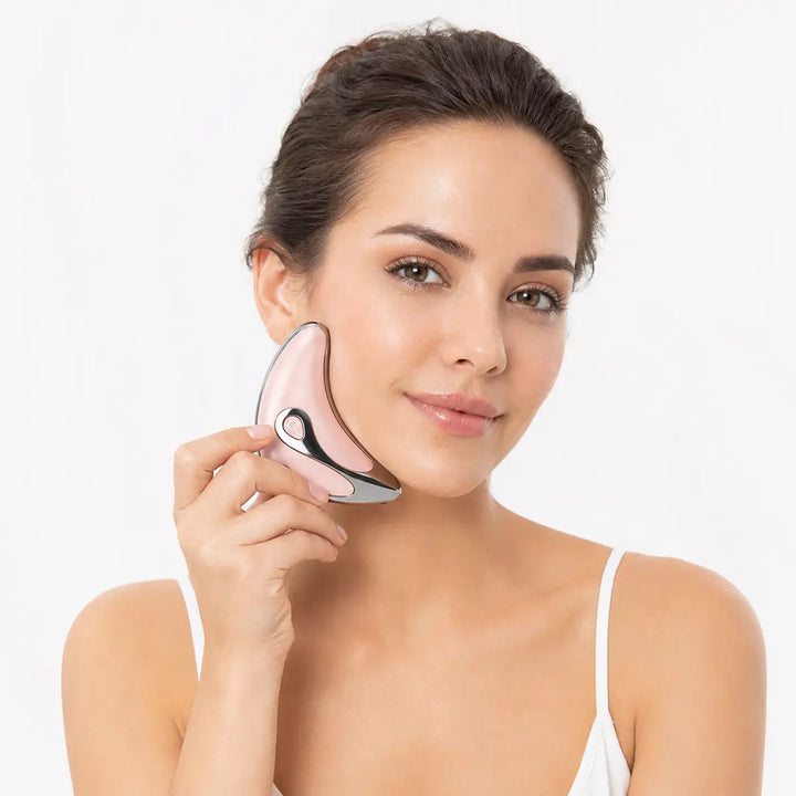 Adult woman, using a pink electric gua sha massager along her cheek and jawline during a clean personal care routine at home.