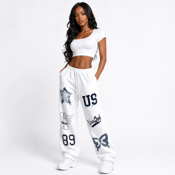 A woman modeling white sweatpants with navy graphic prints, paired with a matching white crop top and white sneakers, against a plain white studio background.