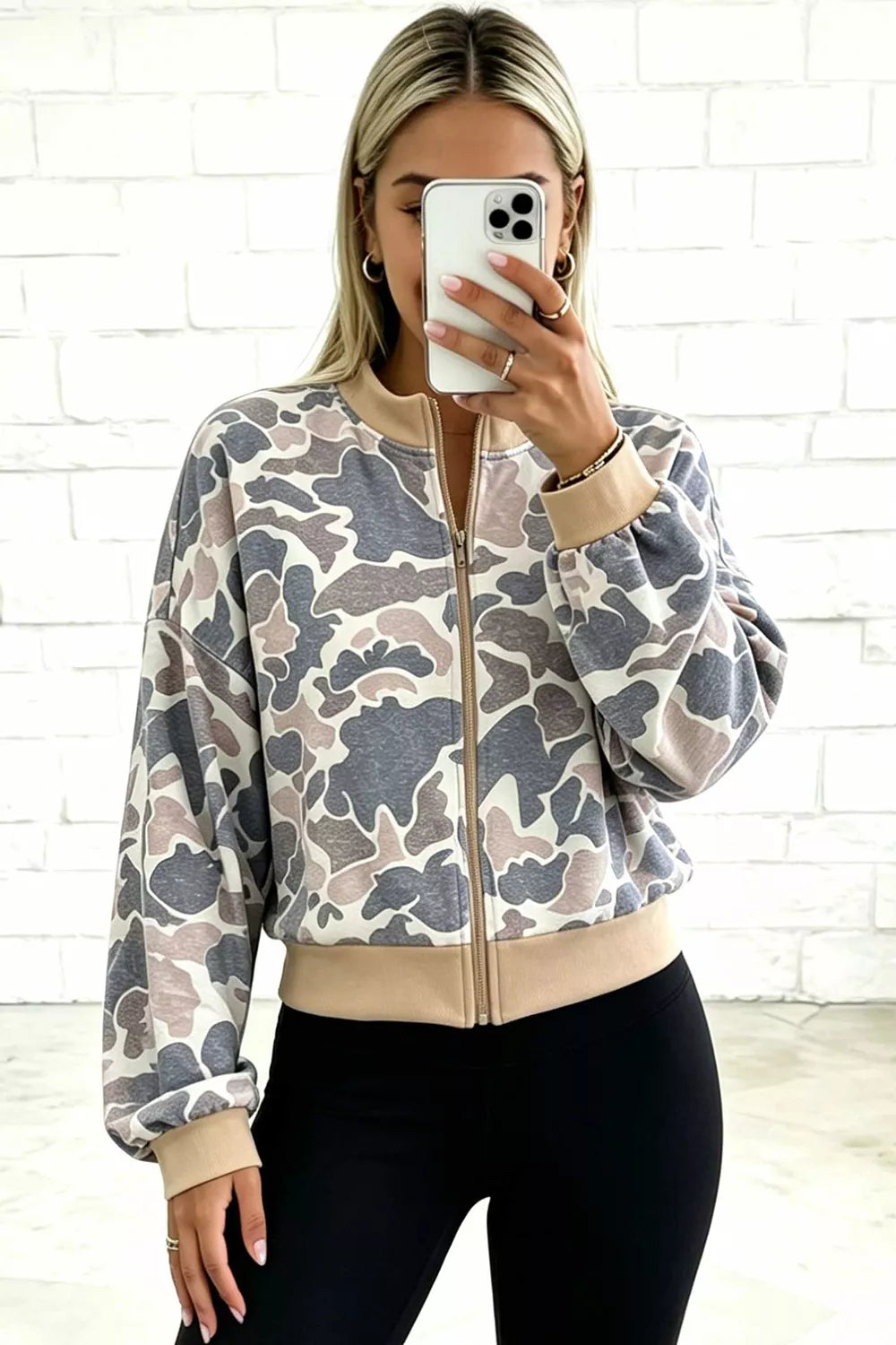 Front view of a model wearing a zipped camouflage bomber with beige ribbed collar, cuffs and hem, styled with black leggings.