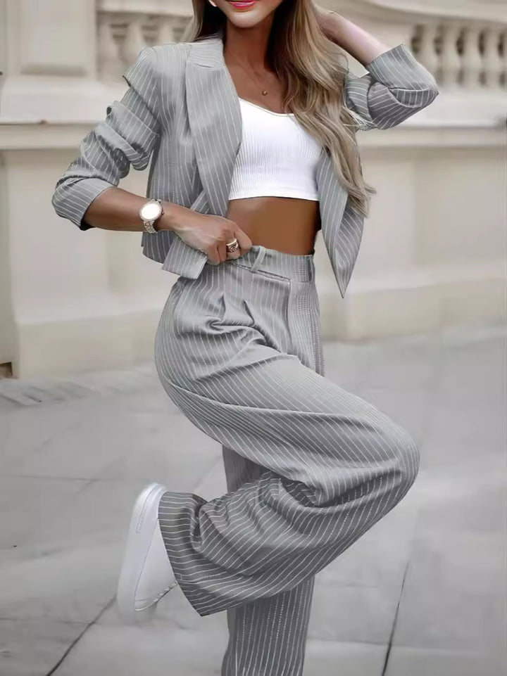 Gray pinstripe cropped jacket and pants shown on an outdoor model in a bent-leg pose, with a white inner top and sneakers.