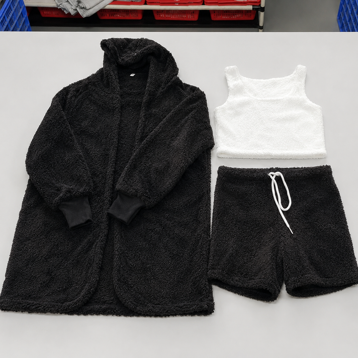 3 piece pajama set women in black with fuzzy hooded cardigan, white crop top and drawstring shorts arranged on a worktable.