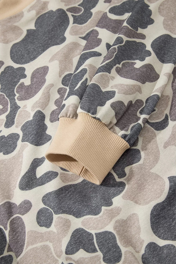 Detail of a camouflage sleeve gathered into a beige ribbed cuff, showing the printed fabric, a seam and finely ribbed finish.