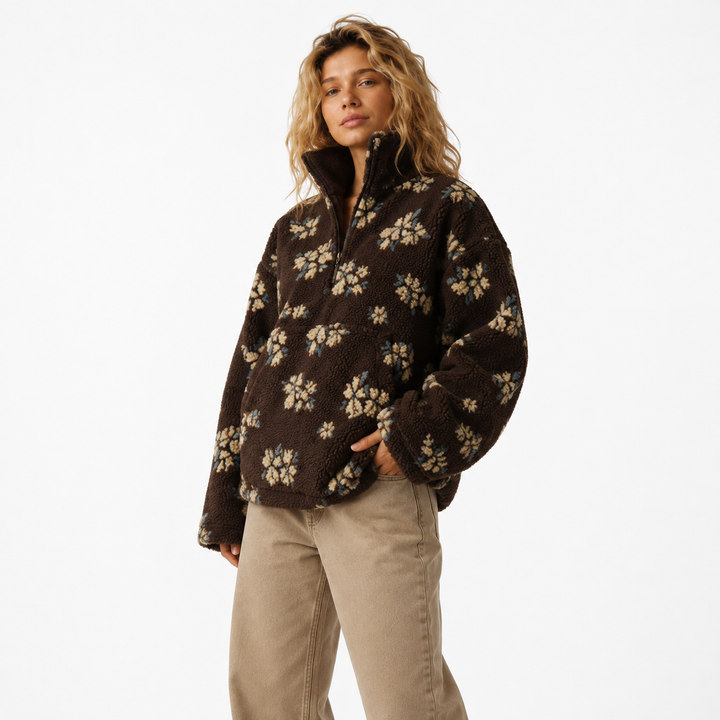 A woman modeling a dark brown fuzzy quarter-zip fleece jacket with a beige floral pattern, paired with beige pants, against a plain white studio background.