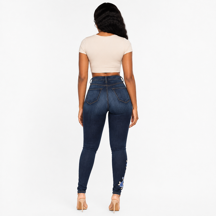 A woman shown from the back wearing dark blue high-waisted skinny jeans with a small floral embellishment near the ankle, paired with a cream crop top and beige heels, against a plain white background.