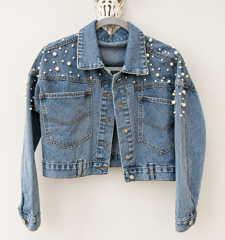 A cropped light blue denim jacket decorated with white faux pearls on the shoulders, chest, and sleeves, hanging on a gray hanger against a light gray background.