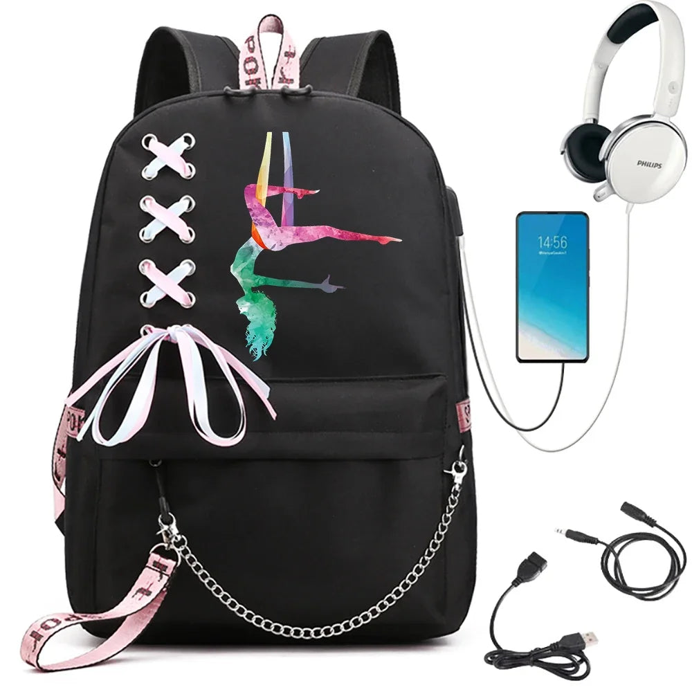 Black backpack featuring a multicolor split gymnast print, pale ribbon bow, pink webbing and chain, beside electronics props.
