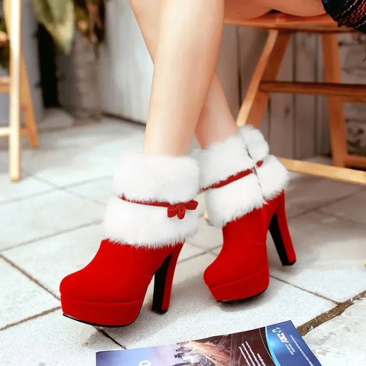 Red boots worn in a seated indoor view, showing the round toes, white fluffy cuffs, side bows, and high heels beside a chair.
