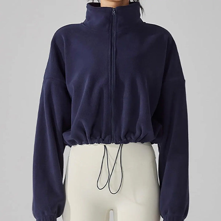 A model wearing a navy blue cropped jacket with a high collar, styled with ivory trousers in front of a gray studio backdrop.
