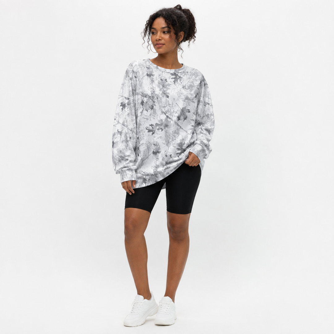 A woman modeling a long-sleeve crewneck sweatshirt with a gray and white abstract floral pattern, paired with black bike shorts and white sneakers, against a plain white studio background.