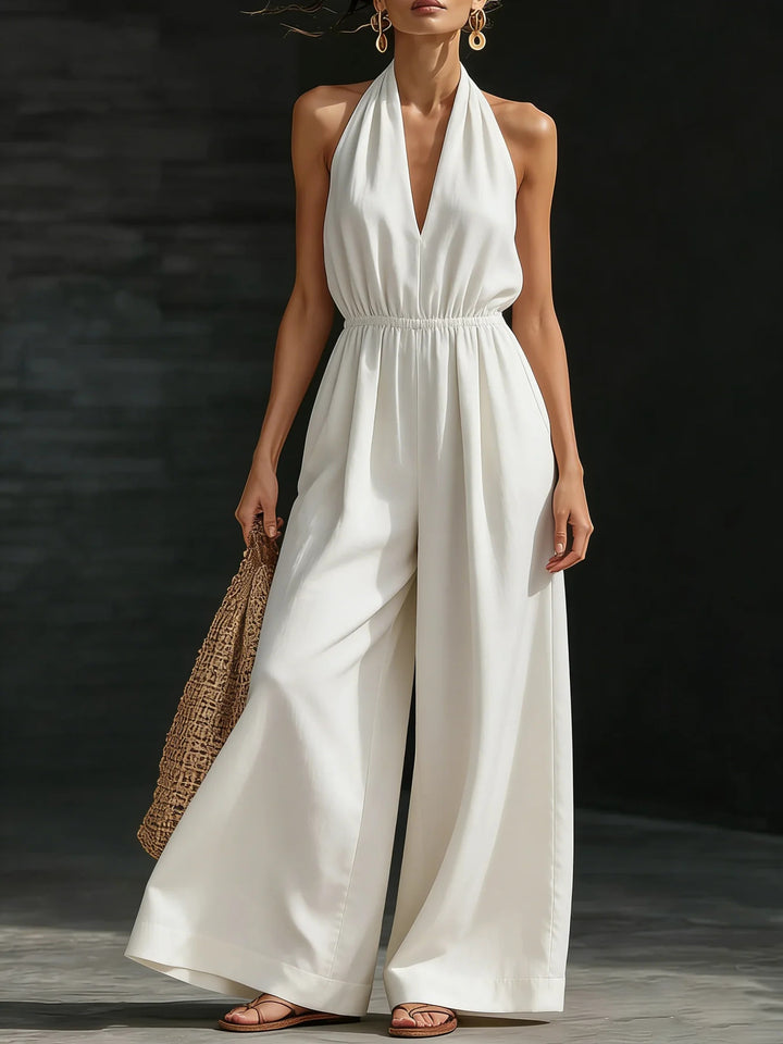 Woman wearing a white halter neck jumpsuit holding a woven bag against a dark background