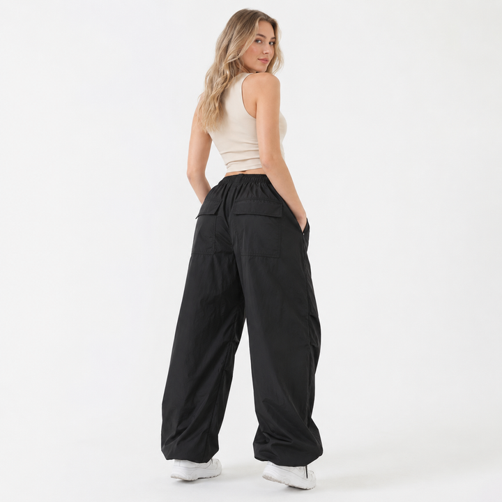 A woman modeling a pair of black wide-leg pants with an elastic waistband and side pockets, paired with a beige crop top and white sneakers, photographed against a solid white studio background.
