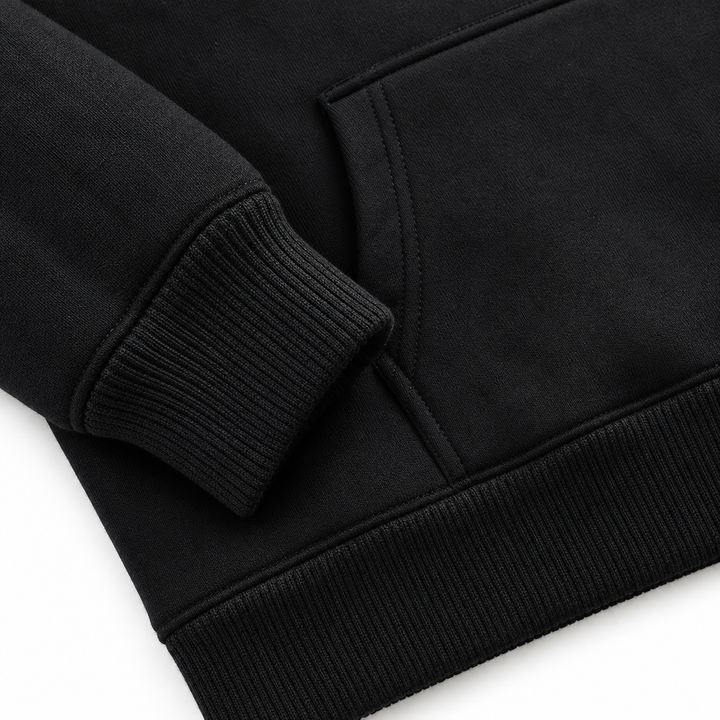 Black pullover hoodie detail showing the ribbed sleeve cuff, kangaroo pocket edge and hem stitching in a bright white studio.