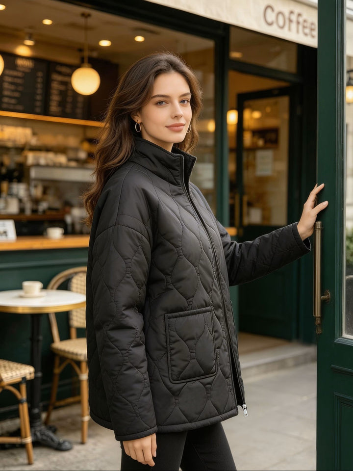 Side view of a woman wearing a black quilted coat outside a cafe showing its stand collar, long sleeves and mid-length shape.