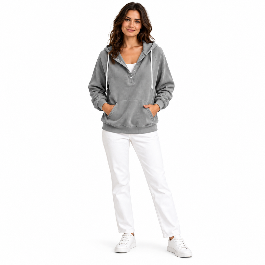 Brunette woman wearing a grey ribbed hooded top with white trousers and sneakers in full-body pose against a studio backdrop.