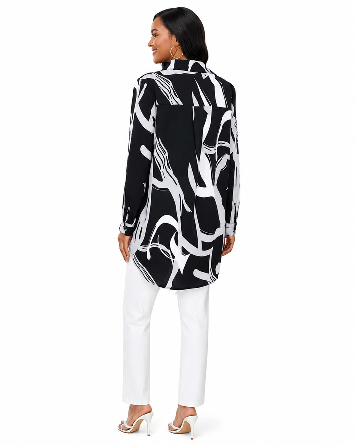 A woman shown from the back wearing a black long-sleeve tunic top with a bold white abstract brushstroke pattern, paired with white pants and white high-heeled sandals, against a plain white background.