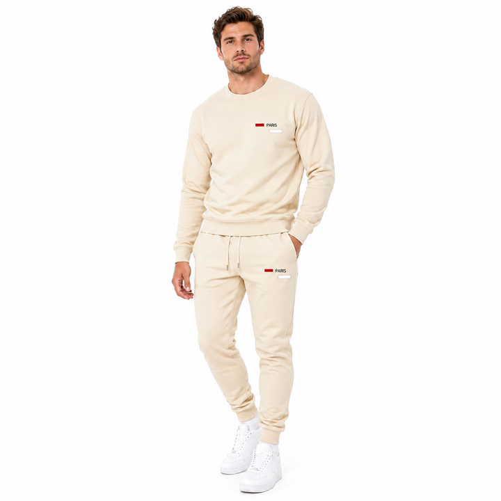 A man modeling a matching beige sweatshirt and jogger pants set with small red and white logo details on the chest and thigh, paired with white sneakers, against a plain white background.