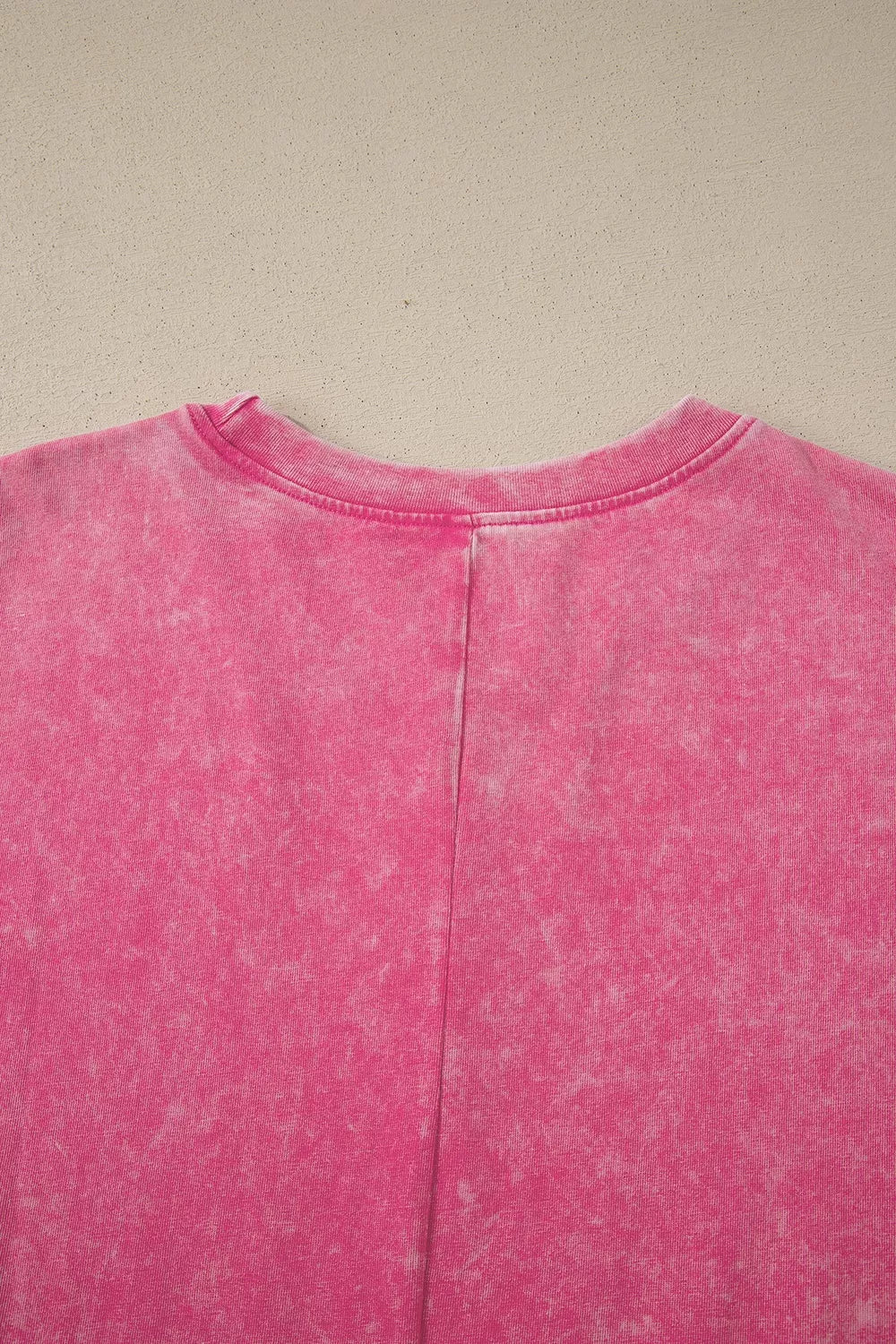 A close-up of a pink acid-wash t-shirt showing the back collar and center seam, laid flat against a neutral beige background.