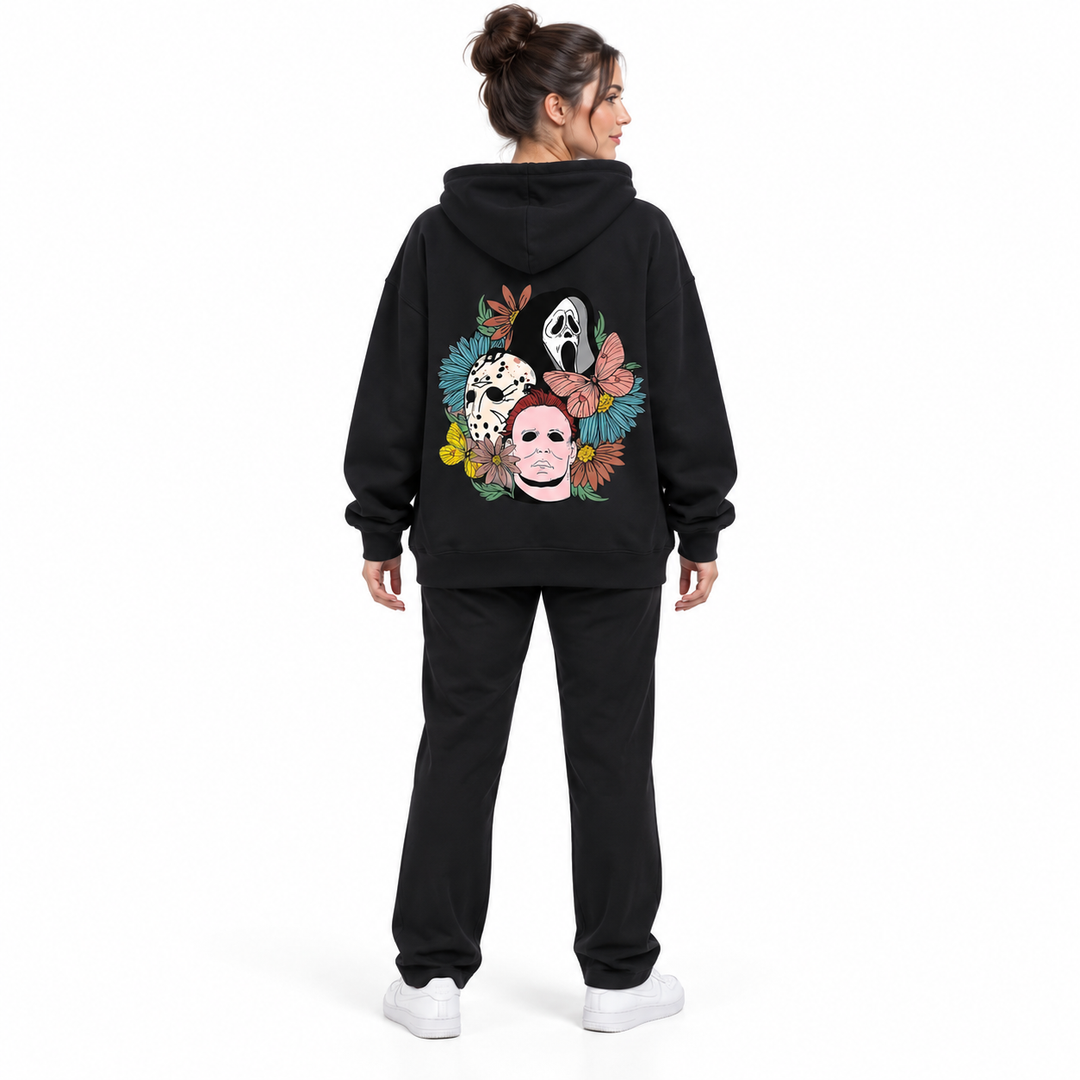 Full-body rear view of a woman wearing the black hooded pullover with floral mask back print, black trousers and white shoes.