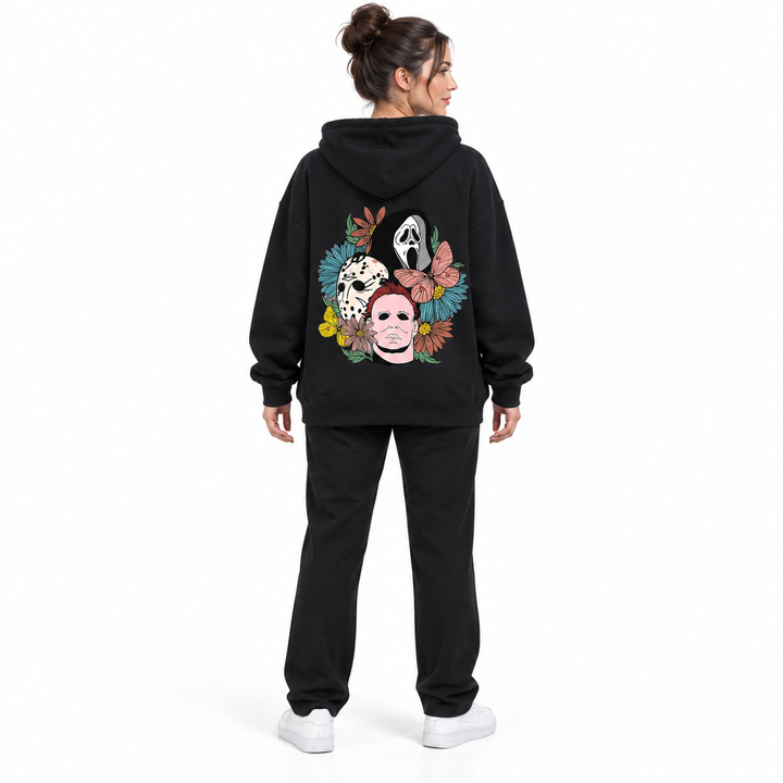 Full-body rear view of a woman wearing the black hooded pullover with floral mask back print, black trousers and white shoes.