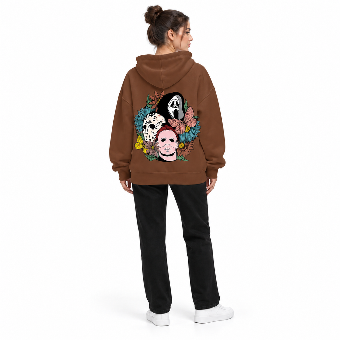 Full-body rear view of a woman wearing the brown hooded pullover with floral mask back print, black trousers and white shoes.