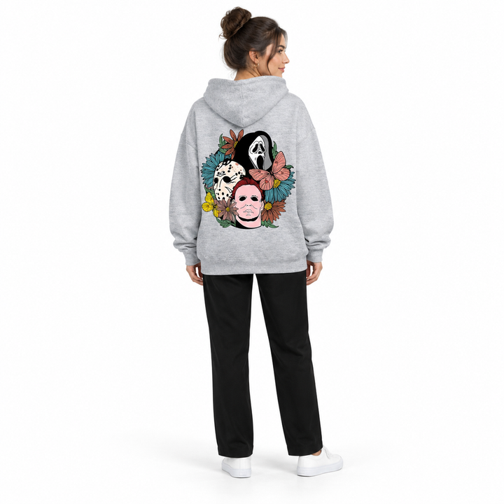 Full-body rear view of a woman wearing the gray hooded pullover with a mask and flower print, black trousers and white shoes.