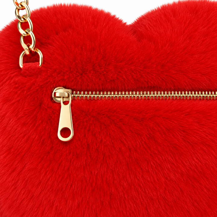 Close-up of a red plush heart bag showing a gold-tone zipper, pull and chain attachment against the red textured bag surface.