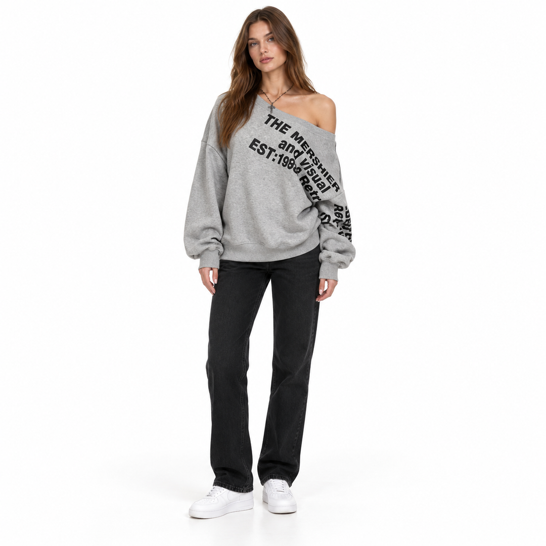 Full-body model in a grey pullover with black lettering, wide neckline, black jeans and white trainers with a white backdrop.