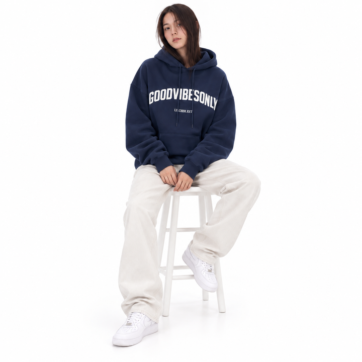 Navy blue hoodie with white chest lettering and a front pocket, worn by a model seated on a white stool with neutral trousers