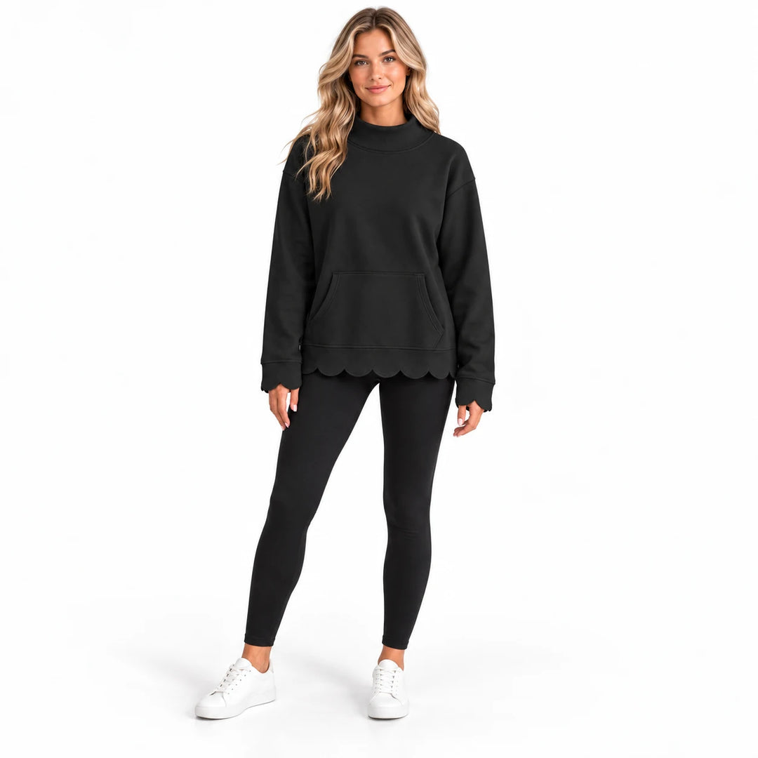 Studio model wearing a black mock neck sweatshirt with scalloped cuffs and hem, styled with black leggings and white sneakers
