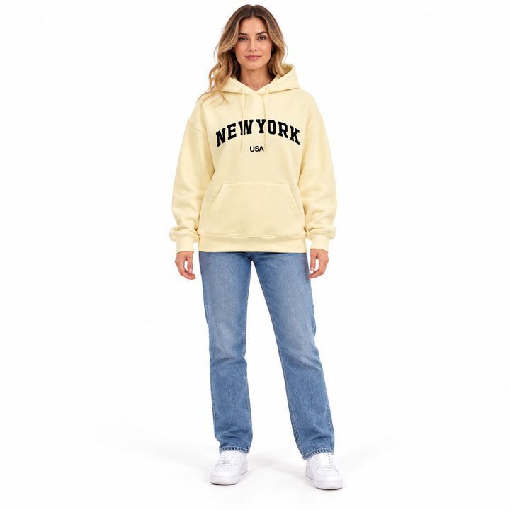 Full body model in a cream hoodie with New York USA graphic print, blue jeans and white sneakers on a plain white background.