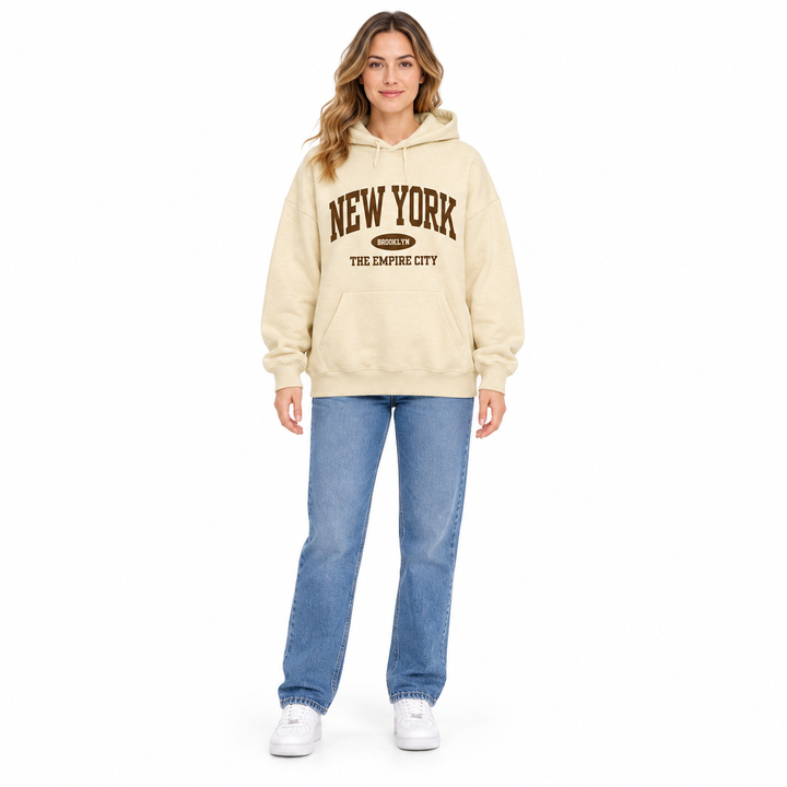 Full body model in a cream hoodie with Empire City chest print, long sleeves, jeans and sneakers on a plain white background.
