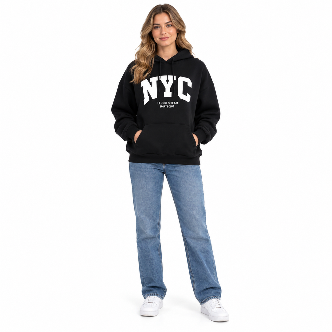 Adult woman wearing a black New York hoodie with white NYC lettering, blue jeans and white sneakers against a white backdrop.