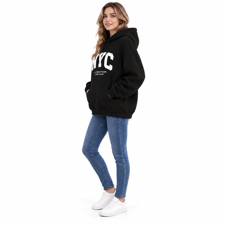 Full body side view of a model in a black hoodie with white NYC print, a front pocket, jeans and sneakers, in a white studio.