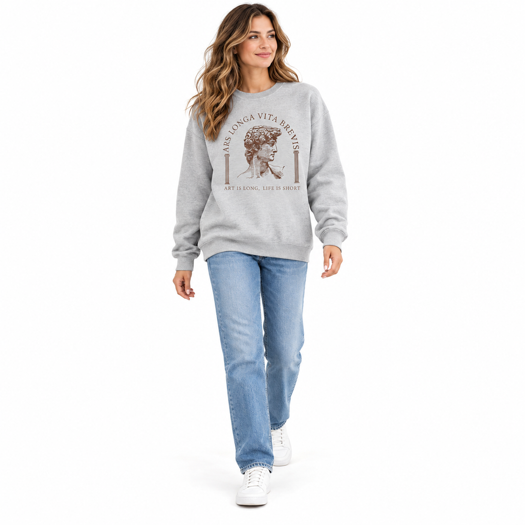 Gray art print sweatshirt on a woman walking in a white studio shown head to shoes with blue jeans and white casual footwear.