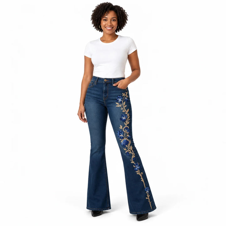 Model wearing dark blue floral flare jeans with blue flowers and gold-tone vines down one leg, in a white studio setting.