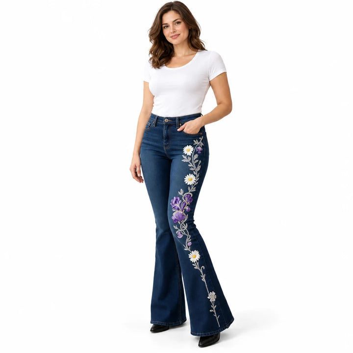 Model wearing dark blue flared jeans with purple irises, white daisies and grey vines printed down one leg, on white.