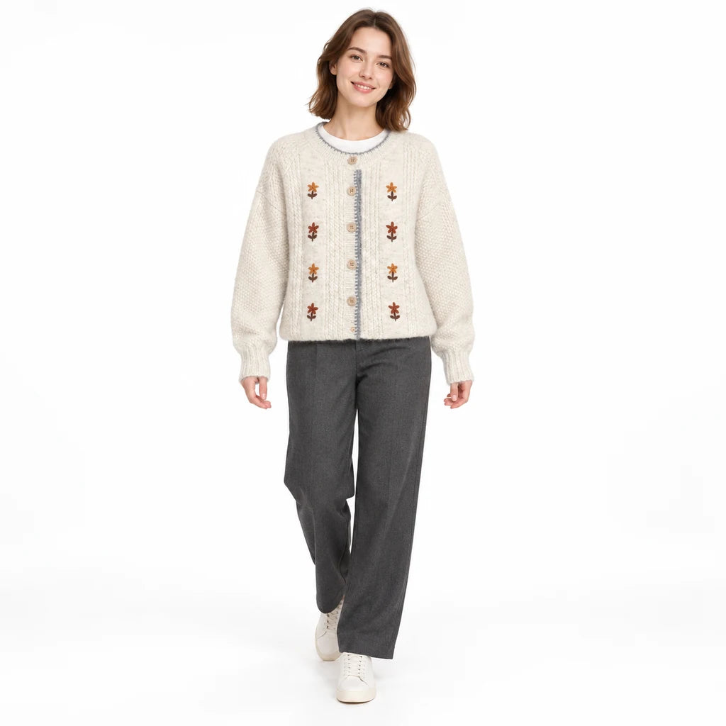 Full-length studio style of an ivory floral cardigan with charcoal trousers and white shoes, as the model takes a small step.