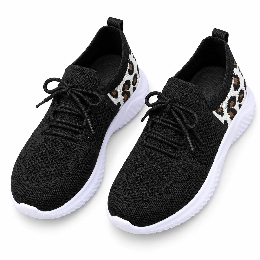 Top view of black lace-up mesh sneakers with round toes, white soles and patterned panels, placed together in a white studio.