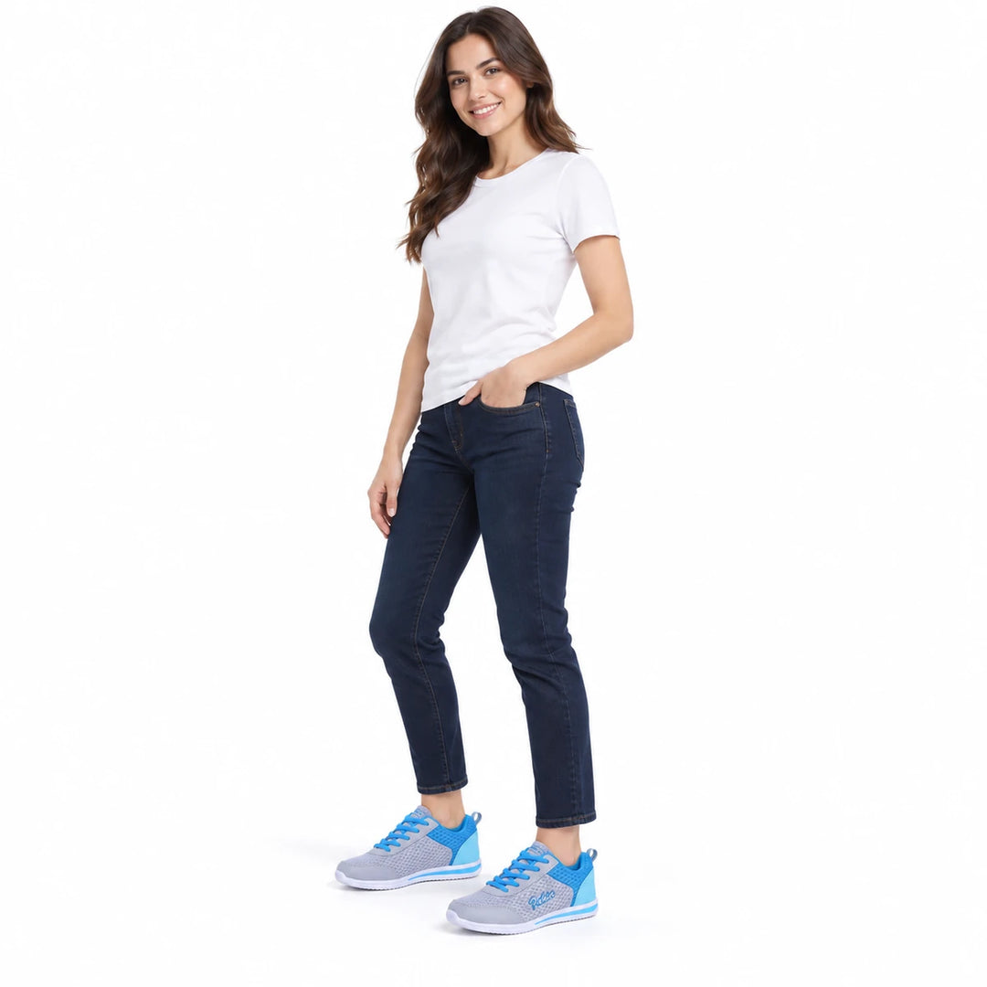 Full-body woman wearing gray and blue lace-up shoes with a white shirt and dark cropped jeans against a white studio backdrop