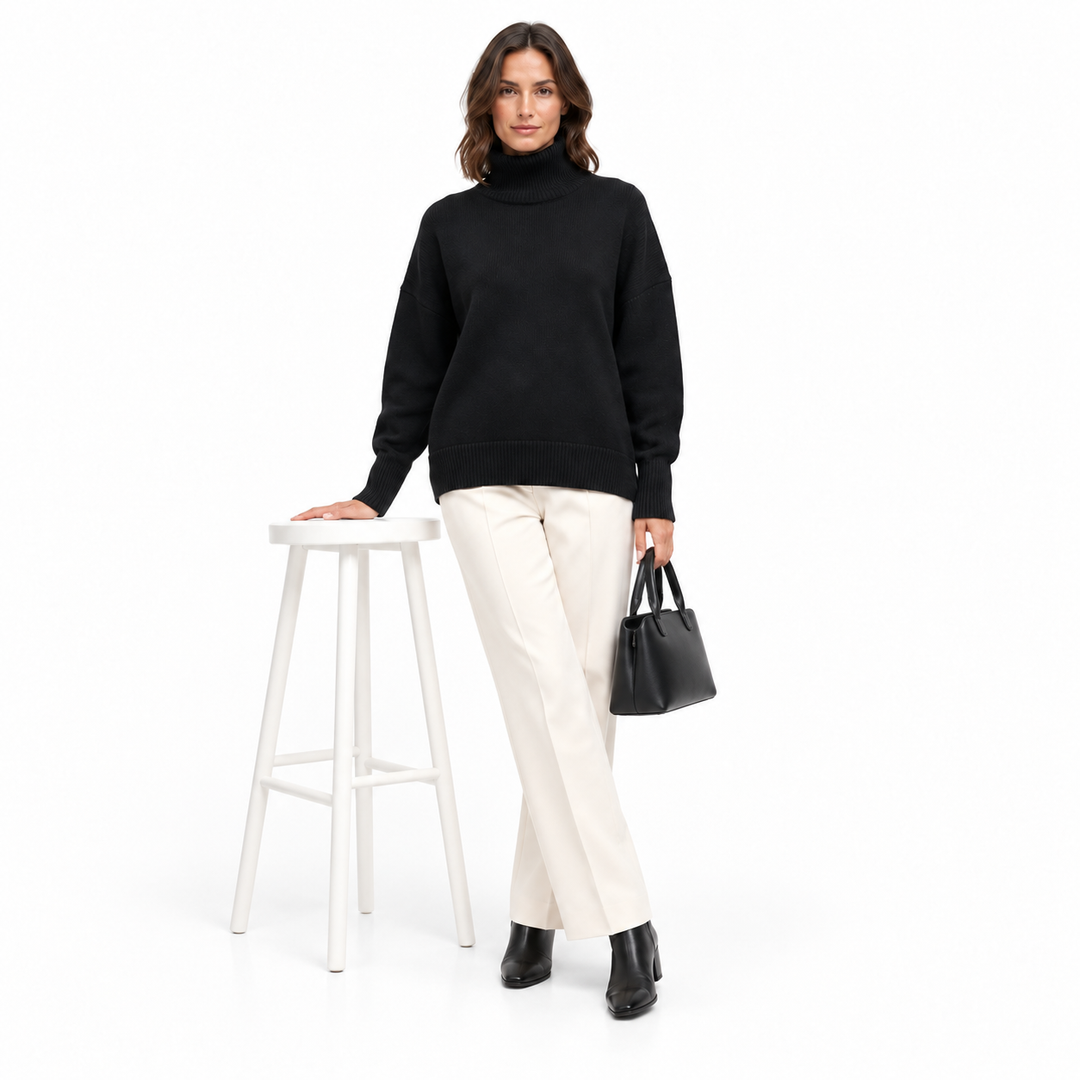 Studio outfit with a model wearing the black turtleneck sweater beside a white stool, carrying a small plain handbag on white