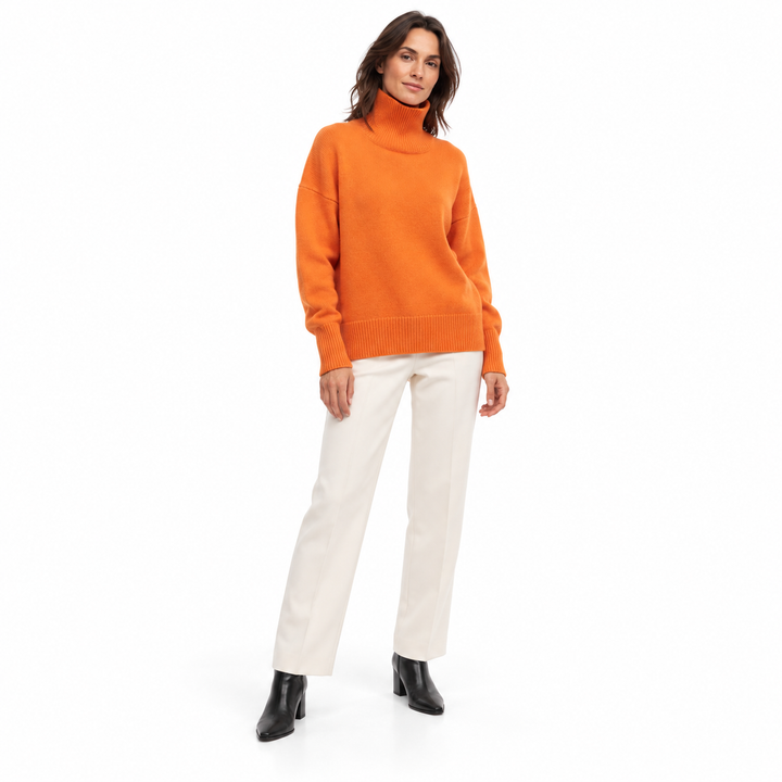 Full-length model wearing an orange red turtleneck sweater styled with ivory tailored trousers and black ankle boots on white
