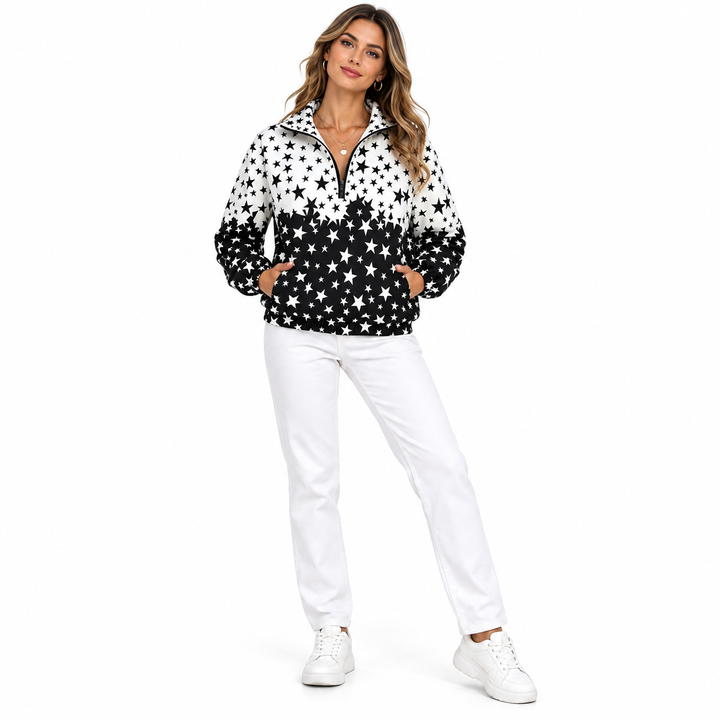 Adult blond woman wearing a black and white star pullover jacket with white trousers and sneakers in a full-body studio view.