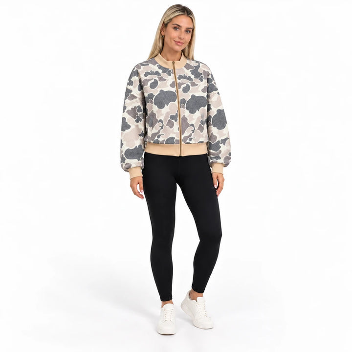 Full-body front view of a blonde model wearing the camouflage bomber with black leggings and white trainers in a white studio