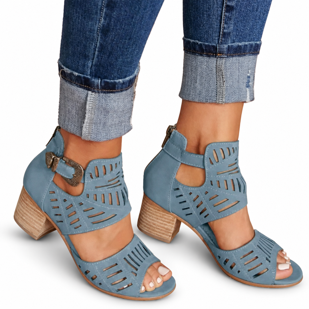 Blue cutout sandals with side buckles, open toes and separate broad heels, worn with cuffed jeans against a white background.
