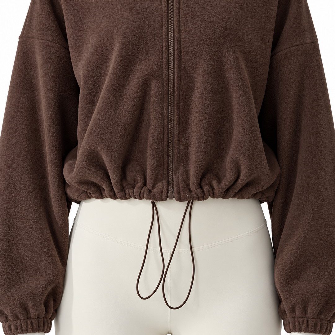 Dark brown jacket detail showing the lower front zipper, gathered cropped hem and long drawcord loops against ivory trousers.