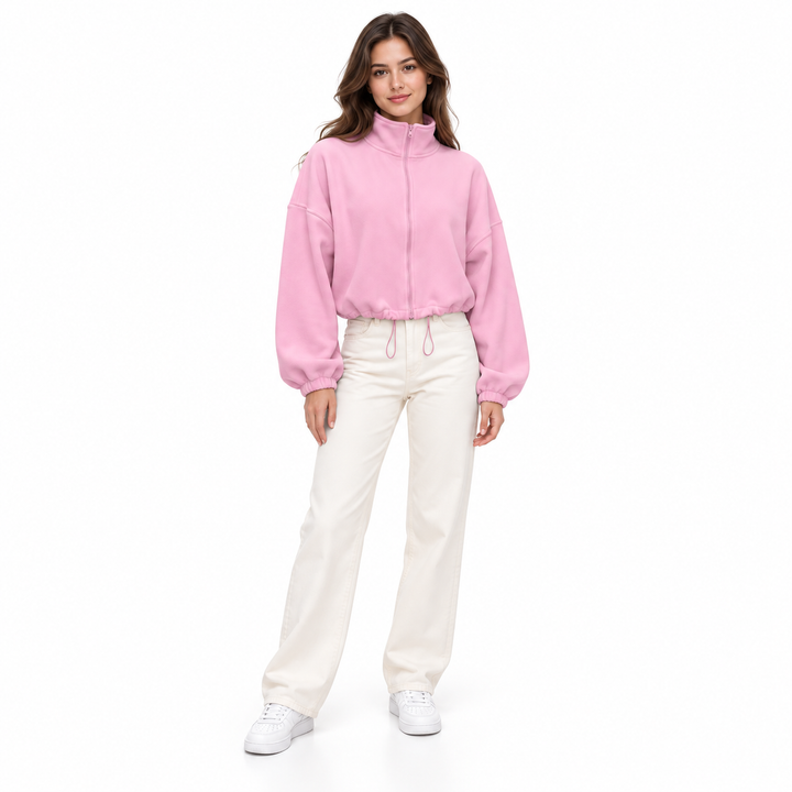 Pastel pink cropped jacket on a full-body brunette model, with stand collar, front zipper, ivory trousers and white sneakers.