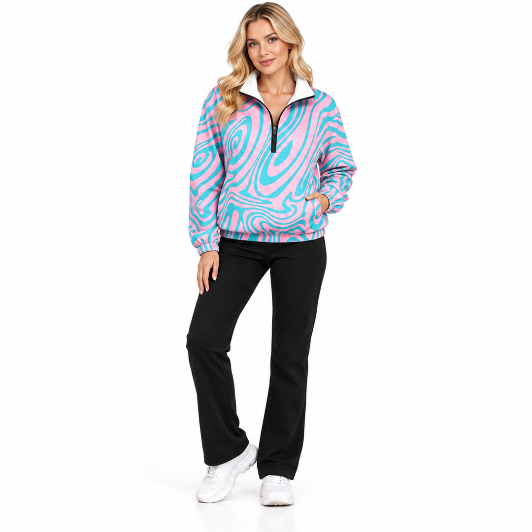 Adult blonde woman wearing a pink and cyan swirl pullover jacket with black trousers and sneakers in a full-body studio pose.