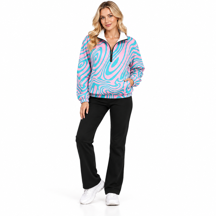 Adult blonde woman wearing a pink and cyan swirl pullover jacket with black trousers and sneakers in a full-body studio pose.