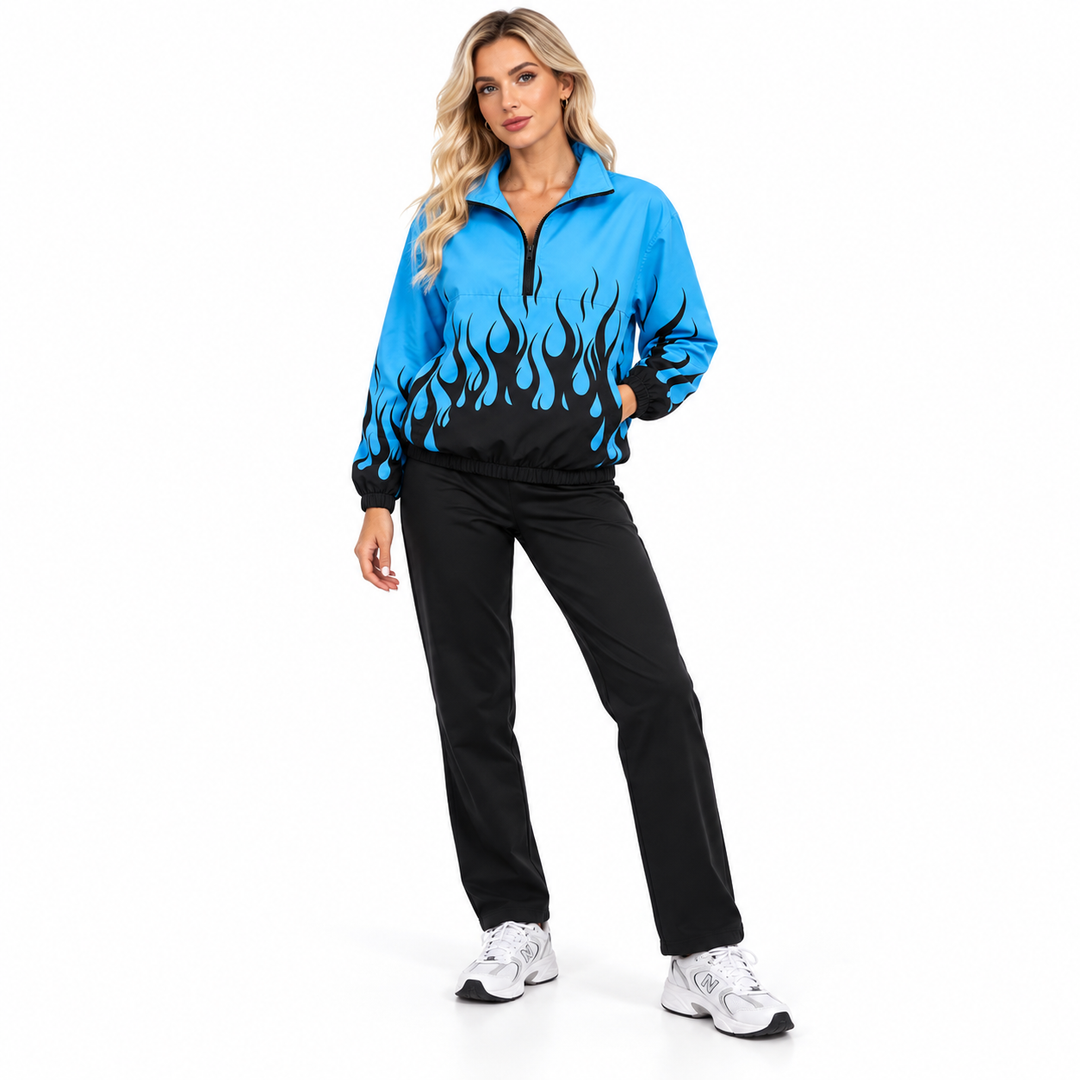Adult blonde woman wearing a blue and dark flame pullover jacket with black trousers and sneakers in a full-body studio pose.