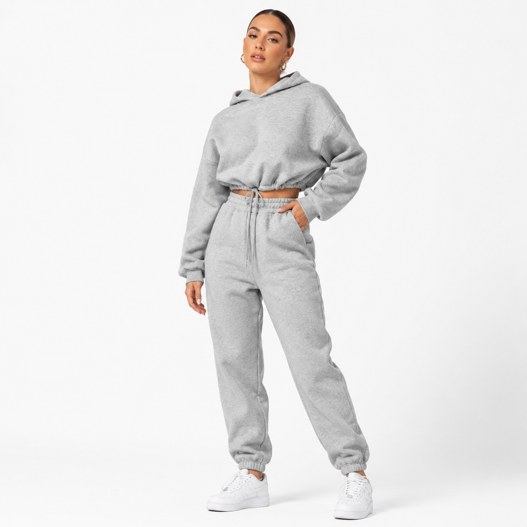 Women tracksuit set in light gray on a female model with cropped hoodie, matching joggers and sneakers on a white studio backdrop.